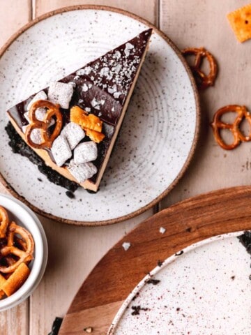A slice of chocolate peanut butter cheesecake on a plate topped with pretzels, cheez its and puppy chow.