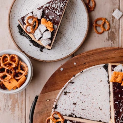 the best chocolate peanut butter cheesecake topped with flaky sea salt, pretzels, puppy chow and Cheez-its