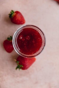Strawberry jam in a glass jar with a few strawberries next to it.