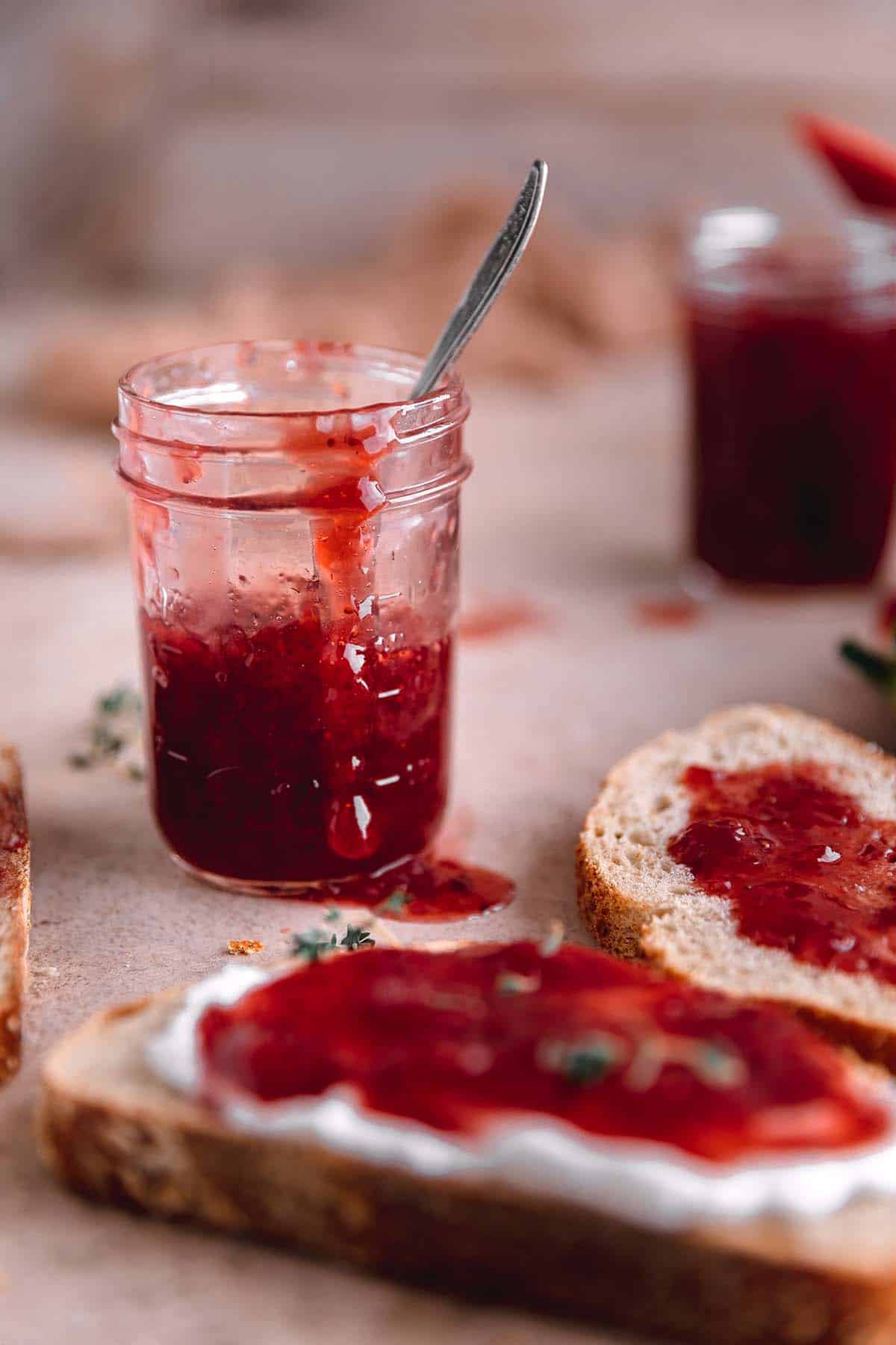 Strawberry Jam in a glass jar with a spoon next to two pieces of toast covered in strawberry jam. 