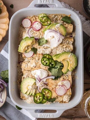 Creamy Green Chile Chicken Enchiladas in a white baking dish topped with sour cream, onion, jalapeno and radish.