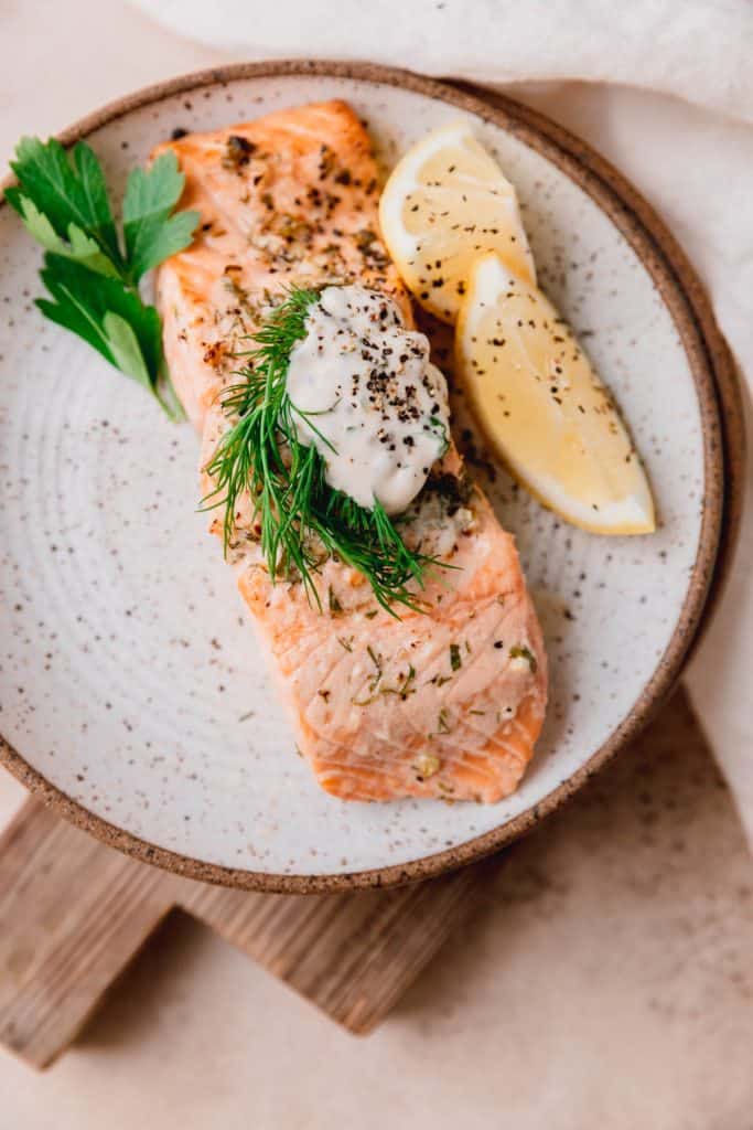 a piece of salmon cooked on a plate with a dollop of tartar sauce, herbs and lemon wedges
