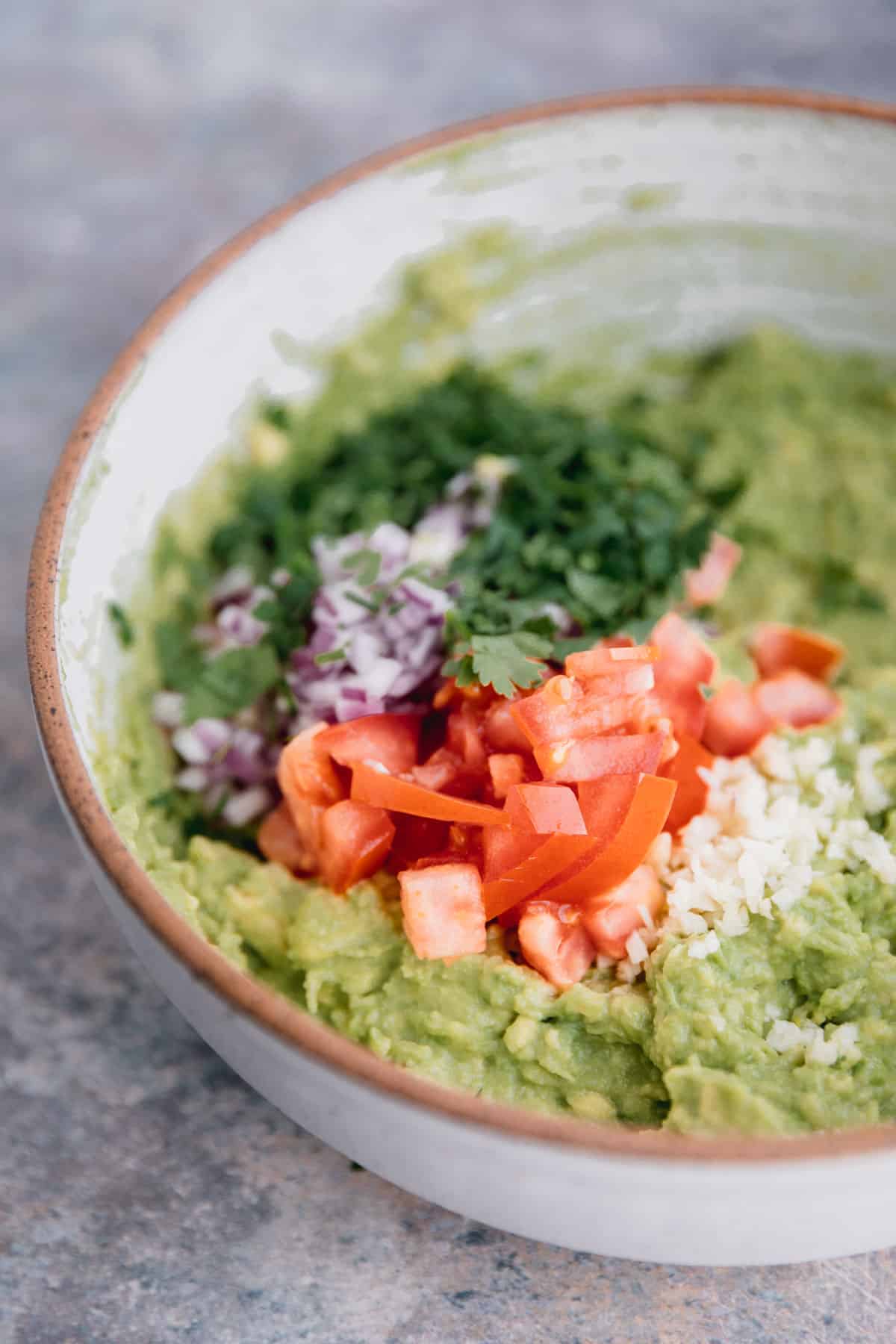 Tomatoes, garlic, onions, cilantro, salt and lime juice being mixed into mashed avocado in a bowl to make guacamole.