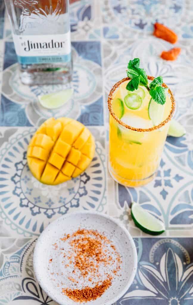 mango chamoy margarita recipe poured over ice with a cut mango, tajin on a small plate and a bottle of el jimador tequila