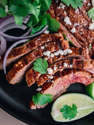 Chipotle Carne Asada sliced on a plate with red onion, cilantro and lime wedges.