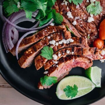 Chipotle Carne Asada sliced on a black plate, topped with sliced red onions, cilantro, queso fresco and lime wedges.