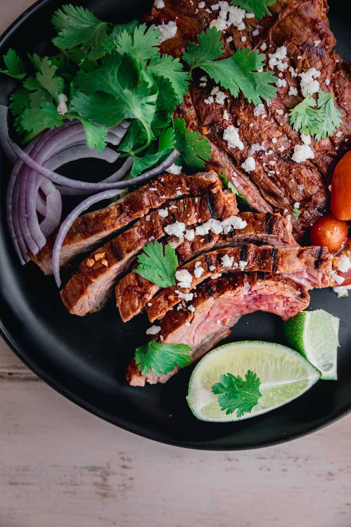 Chipotle Carne Asada sliced on a black plate, topped with sliced red onions, cilantro, queso fresco and lime wedges. 