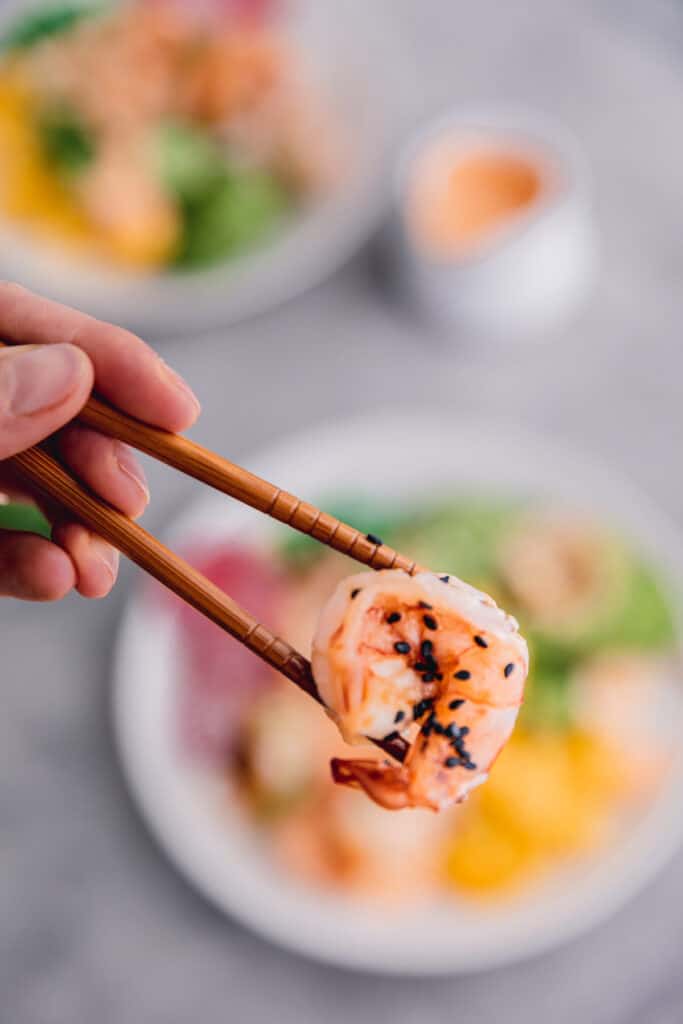 a piece of marinated shrimp with black sesame seeds being held up over the bowl with chopsticks
