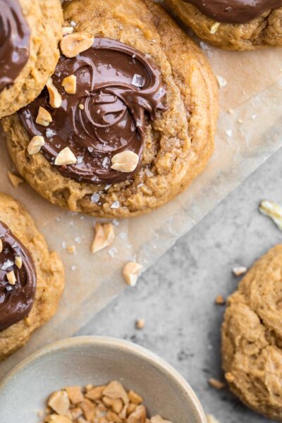 multiple pb2 cookies with swirled Nutella tops and crushed peanuts with flaky sea salt on top of the Nutella. The cookies are sitting on a sheet of parchment paper with a small dish of peanuts to the side