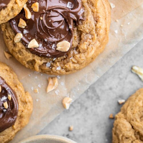 multiple pb2 cookies with swirled Nutella tops and crushed peanuts with flaky sea salt on top of the Nutella. The cookies are sitting on a sheet of parchment paper with a small dish of peanuts to the side