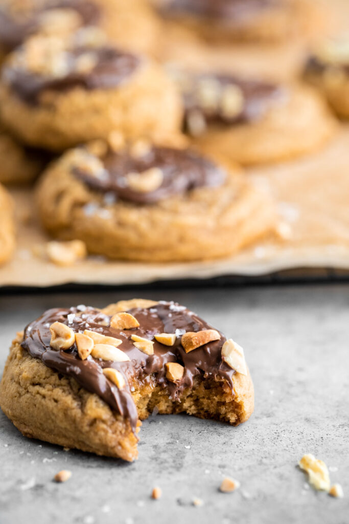 a peanut butter cookie topped with Nutella, peanuts and flaky sea salt with a bite out of it. More cookies on a parchment paper lined cooling rack behind the single cookie.