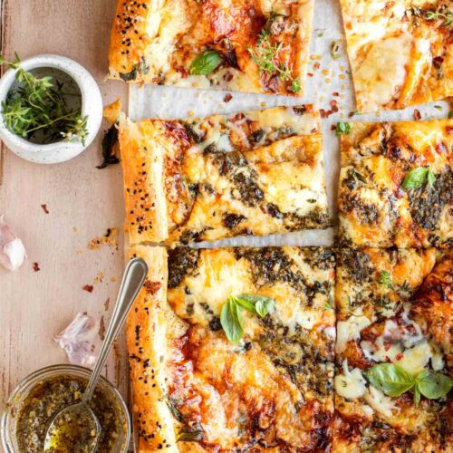 A puff pastry cheese tart cut into rectangles sitting on top of parchment paper that's on a wooden surface. Garnished with basil leaves and has pesto, thyme and honey on the side in small dishes.
