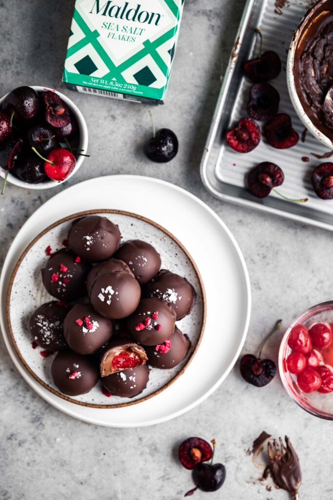 A couple white plates with chocolate covered cherries, a box of Maldon's sea salt, a sheet pan with a bowl of chocolate, one bowl of fresh cherries and a bowl of maraschino cherries.