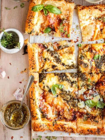 A puff pastry cheese tart cut into rectangles sitting on top of parchment paper that's on a wooden surface. Garnished with basil leaves and has pesto, thyme and honey on the side in small dishes.
