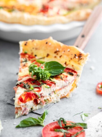 A slice of tomato pie on a spatula, garnished with a whole basil leaf. The rest of the tomato pie is in a dish just behind the slice.