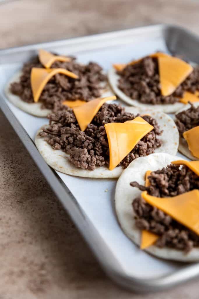 Tortillas with ground beef and American cheese triangles on a sheet tray. 