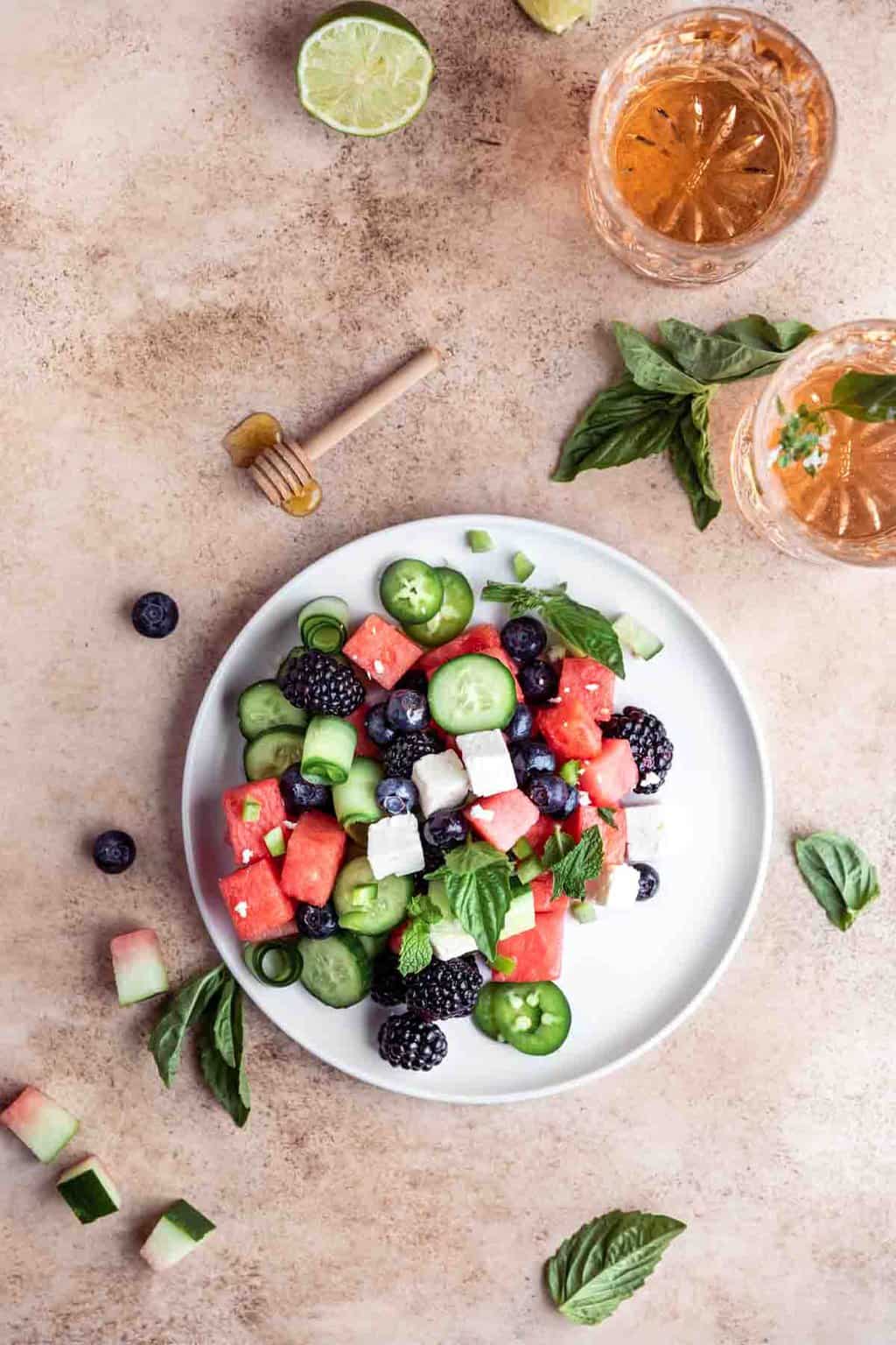 Watermelon feta salad with berries on a plate alongside a small dish of honey and cut limes. 