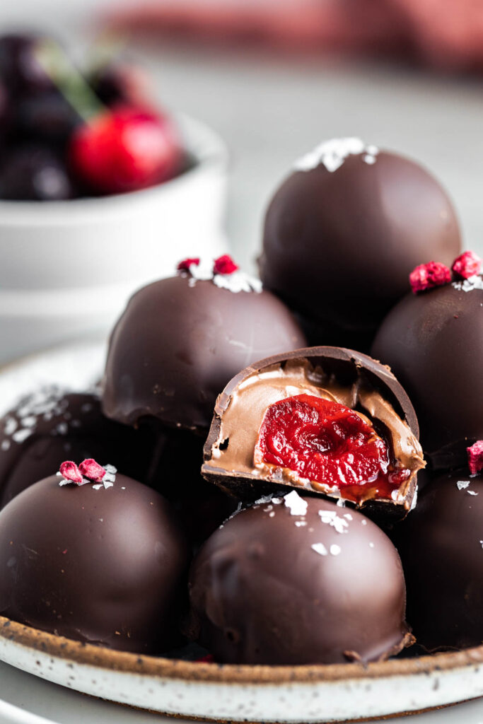 a pile of chocolate covered cherries on a plate with a bite taken out of one so you can see the Nutella and cherry in the center. A white bowl with fresh cherries behind the plate.