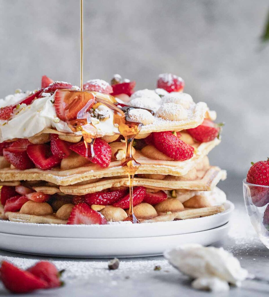 A stack of bubble waffles on a white plate topped with honey lemon cream, and strawberries. A stream of syrup being poured on top and running down the sides.