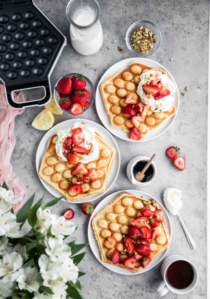 3 plates of bubble waffles topped with strawberries, syrup and honey lemon cream. A waffle machine off to the side. 