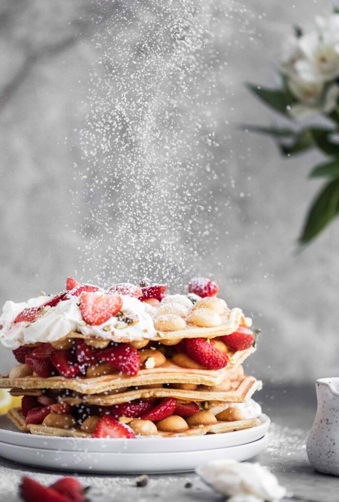 Powdered sugar being dusted on top of the waffles that are topped with strawberries and cream on a plate.