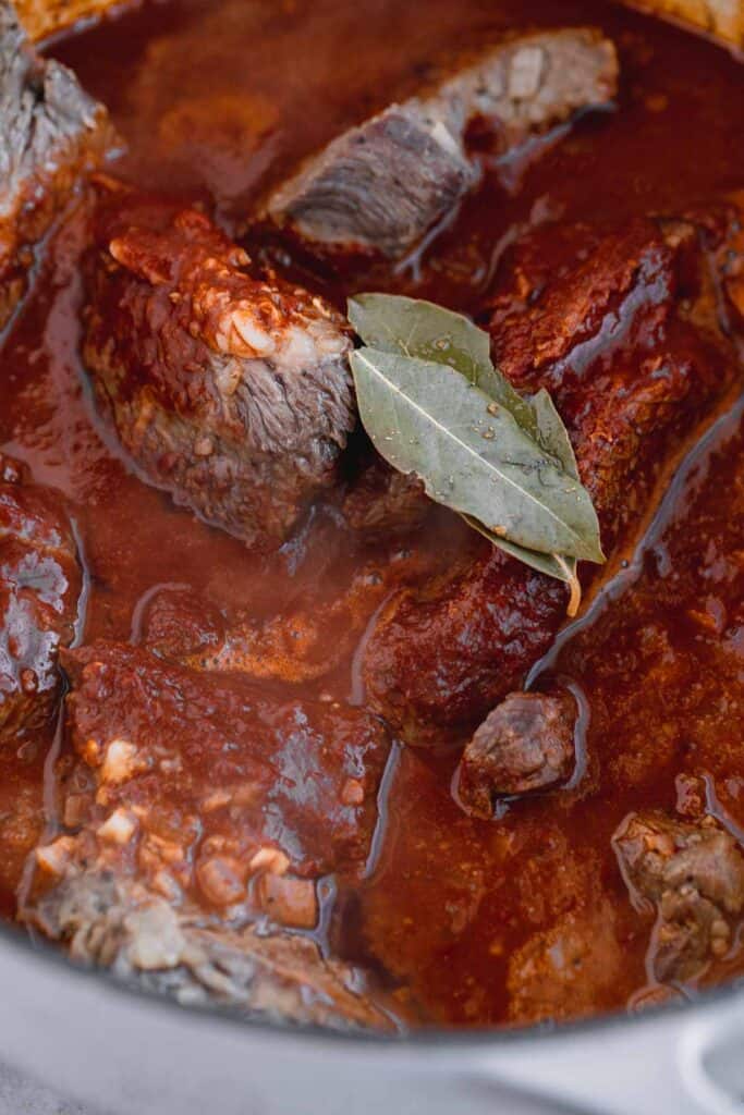 beef chuck roast being braised in a Dutch oven with dried chile sauce, spices, onion, garlic and bay leaves