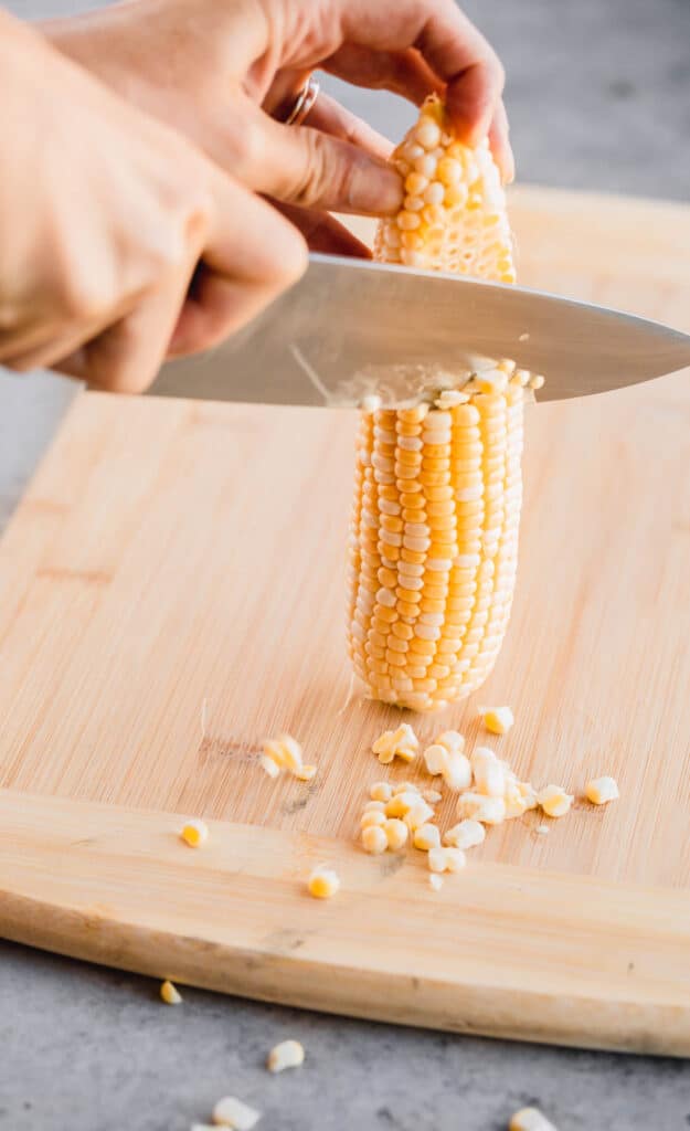 corn being cut off the cob with a chef's knife on a wooden cutting board