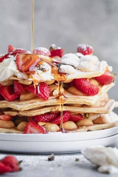 A stack of bubble waffles on a white plate topped with honey lemon cream, and strawberries. A stream of syrup being poured on top and running down the sides.