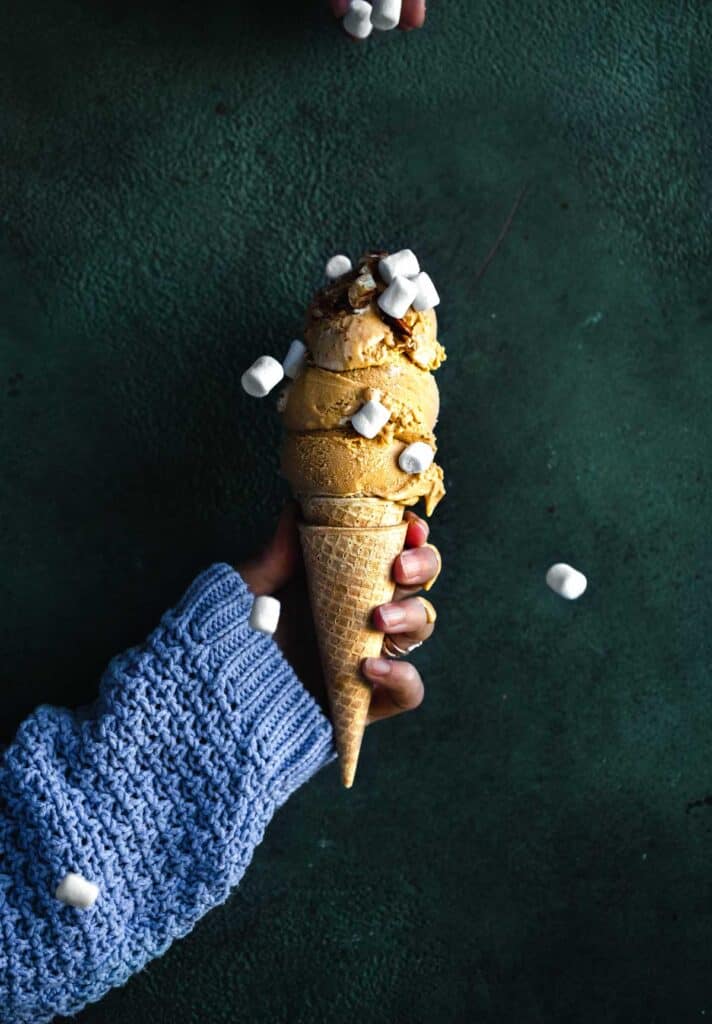 Three scoops of sweet potato ice cream on a sugar cone that is topped with pecans. The cone is being held by a single hand that is wearing a purple sweater. Mini Marshmallows are being dropped on top of the cone from above.