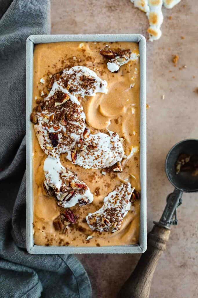 Sweet potato ice cream frozen in a loaf pan that's been swirled with toasted marshmallow, pecan pieces, brown sugar and cinnamon. A grey linen napkin lying to the left of the loaf pan, an ice cream scoop to the right and toasted mini marshmallows above the pan.