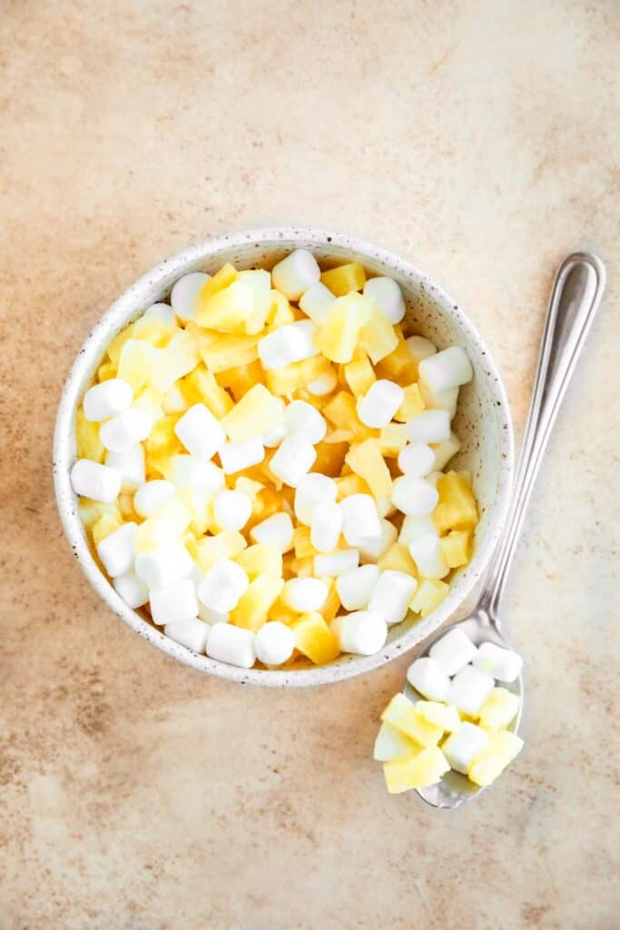 Marshmallow and pineapple mixed together in a small bowl with a spoon full of the mixture set to the side.