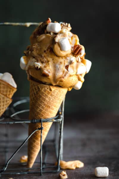 An ice cream cone with two scoops of homemade sweet potato ice cream on top of it that's topped with marshmallows and pecans. The cone is standing upright in a metal stand.