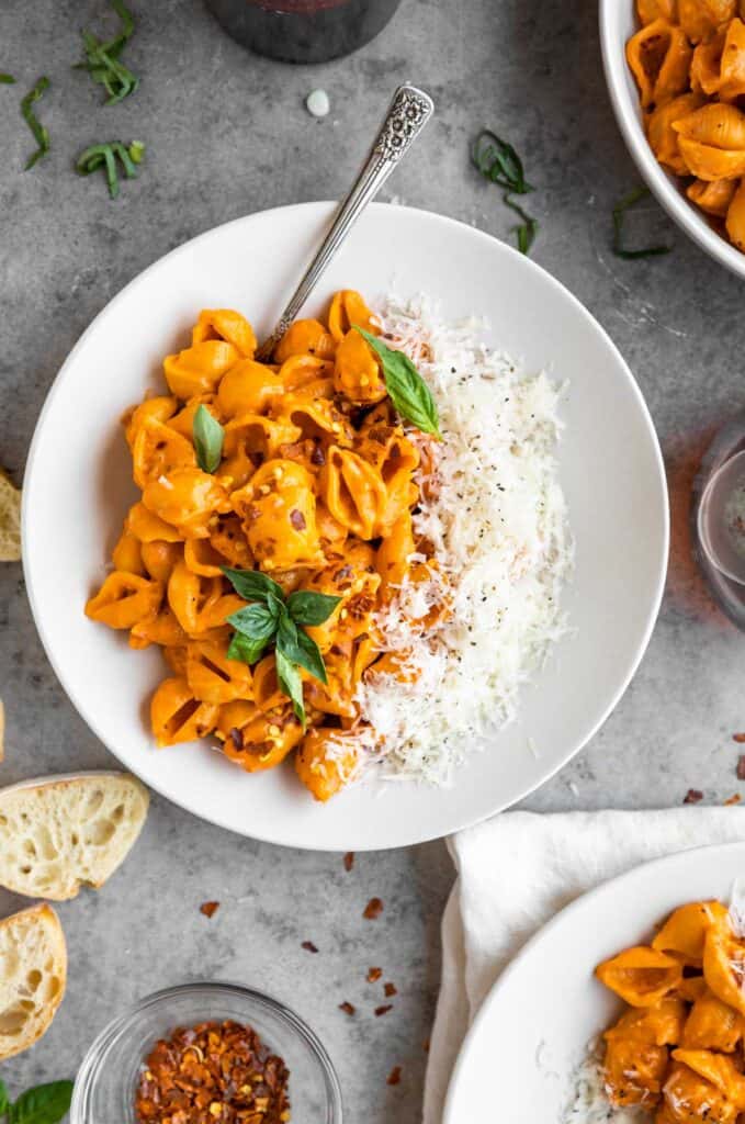 Gigi Hadid pasta recipe in a bowl topped with cheese and fresh basil. Two more bowls of pasta to the side with sliced baguette and crushed red pepper in a small round dish.
