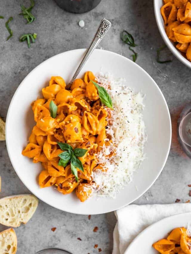 Gigi Hadid pasta recipe in a bowl topped with cheese and fresh basil. Two more bowls of pasta to the side with sliced baguette and crushed red pepper in a small round dish.