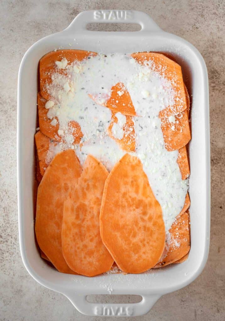 Sweet potato slices being layered into a white casserole dish, alternating directions each later. The potato slices are topped with the cream mixture and Romano cheese.