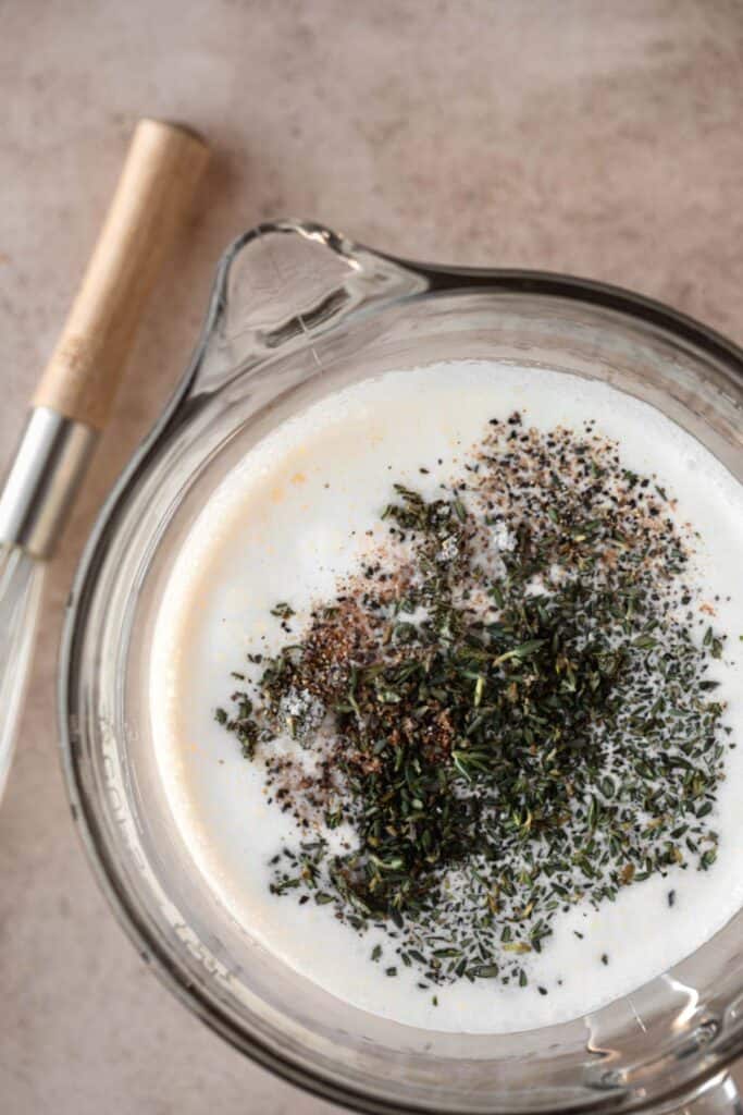 Cream mixture in a clear glass pitcher with the herbs, garlic and seasonings sitting on top of it and a whisk lying to the side.