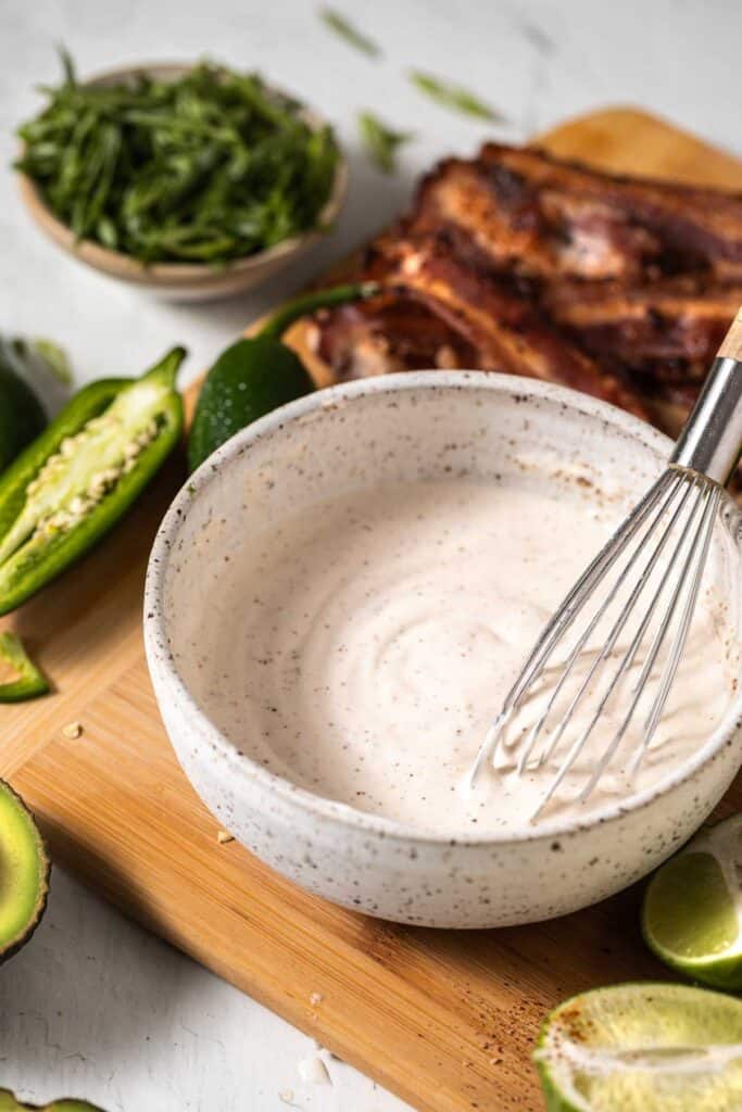 Creamy lime dressing whisked together in a small white bowl with jalapenos, green onion, bacon and limes surrounding the bowl.