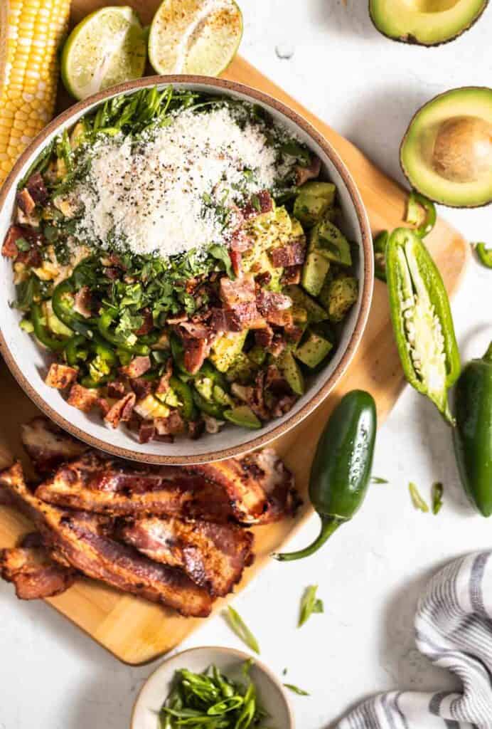 Corn salad in a bowl without the dressing. Bacon, jalapenos, avocado and limes lying around the bowl.