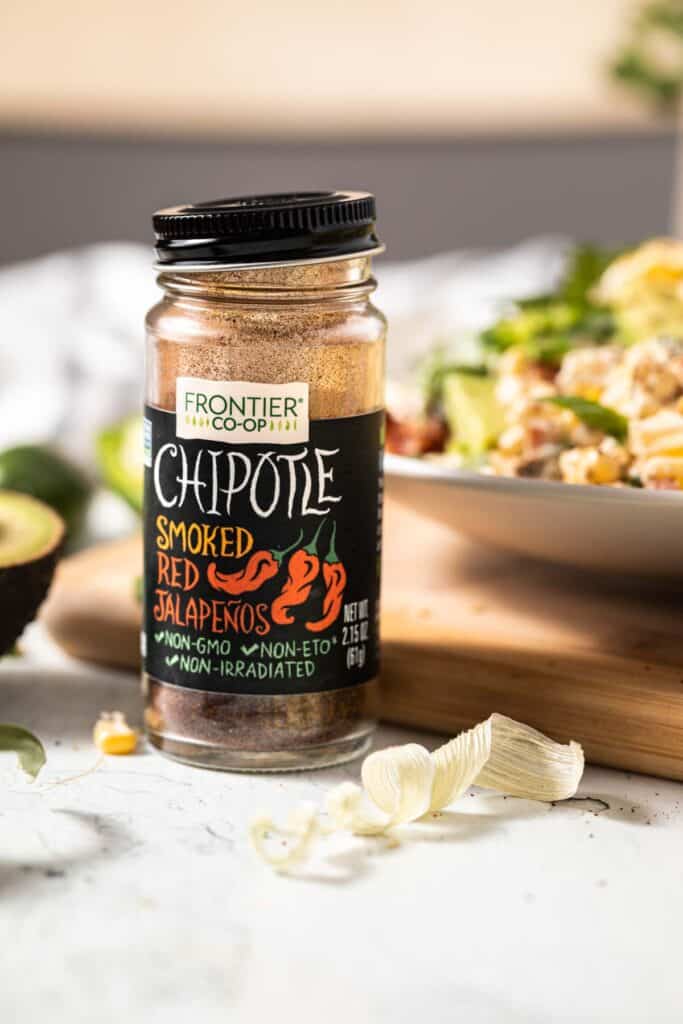 A bottle of chipotle chile powder in front of a cutting board that has a bowl of crack corn salad on it. 