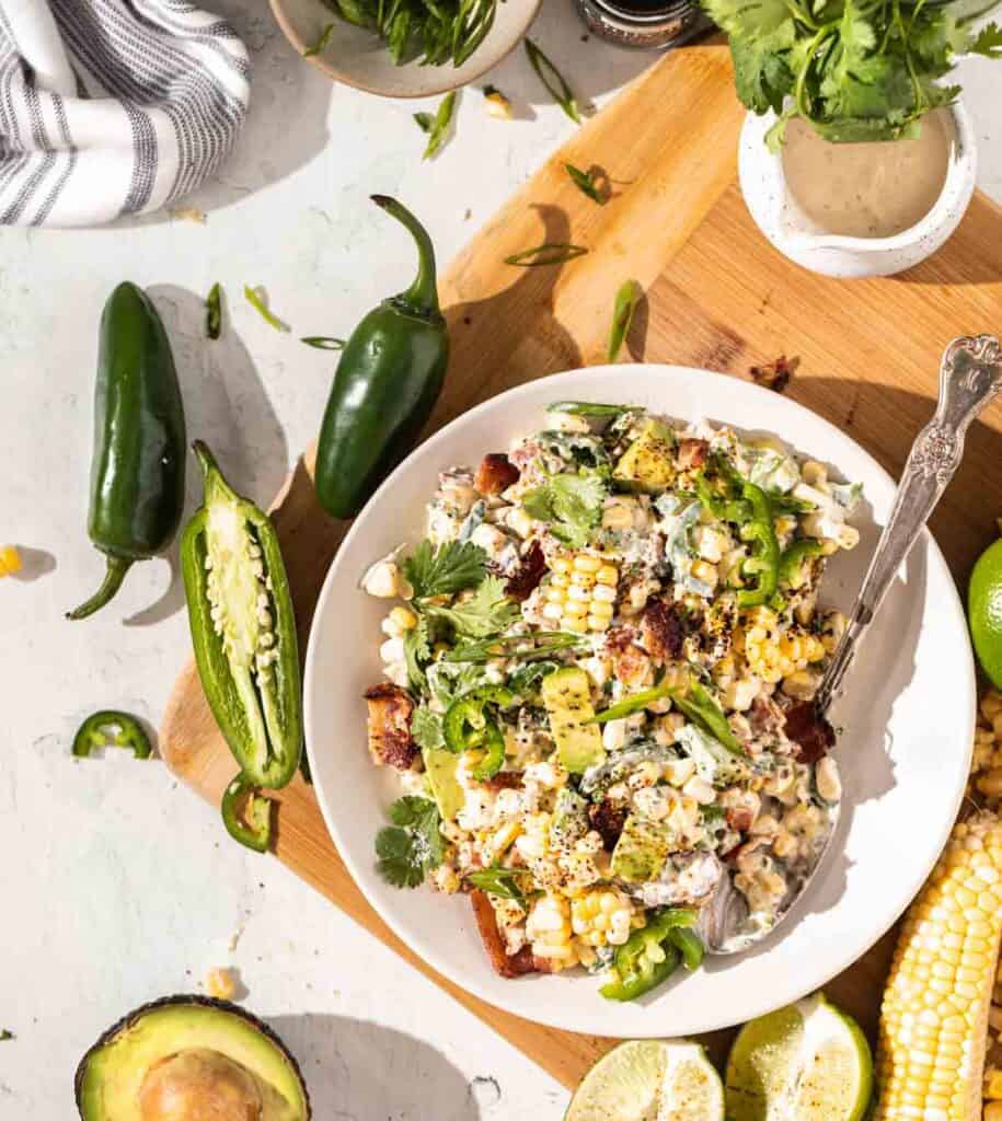 Crack Corn Salad in a white bowl on top of a light wood cutting board. A metal spoon in the bowl of corn with ingredients scattered around.