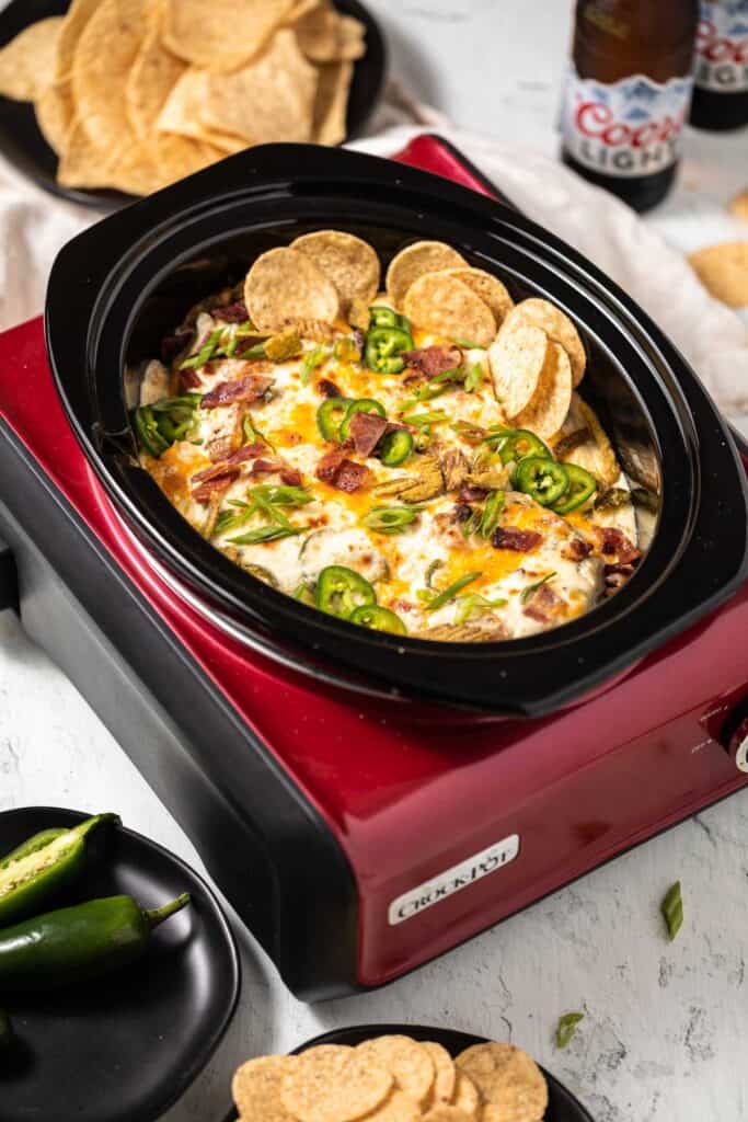 Jalapeno Popper Dip in a red crock pot with a black insert. A couple bottles of beer, tortilla chips and whole jalapenos on black plates to the side of the crock pot. 