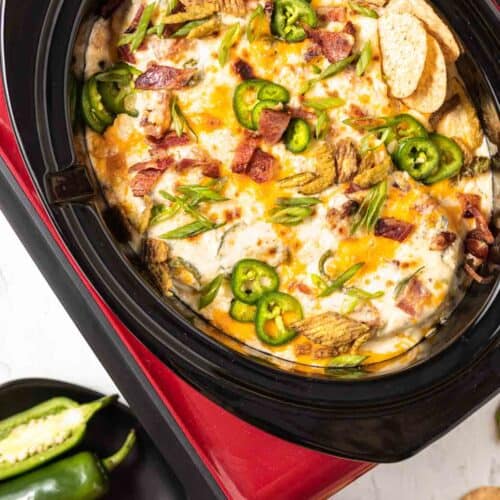 A red crockpot with a black insert filled with cooked jalapeno popper dip and topped with bacon, green onion, crispy jalapenos and round tortilla chips. Black plates of jalapenos and tortilla chips to the side.
