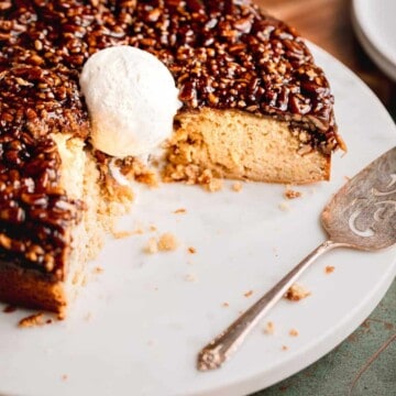 Pecan Upside Down Cake on a white marble circular tray with ⅓ of the cake cut out. One scoop of vanilla ice cream on top of the cake and a serving spatula next to it.
