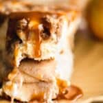 A slice of cheesecake stuffed with apple pie filling that has been topped with oat streusel and caramel sauce sitting on top of a yellow decorative plate with one bite taken off the end.