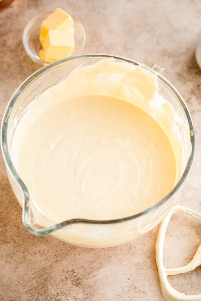 Cheesecake mixture blended together in the clear glass bowl of a stand mixer with the paddle attachment to the side as well as a dish of cut butter. 