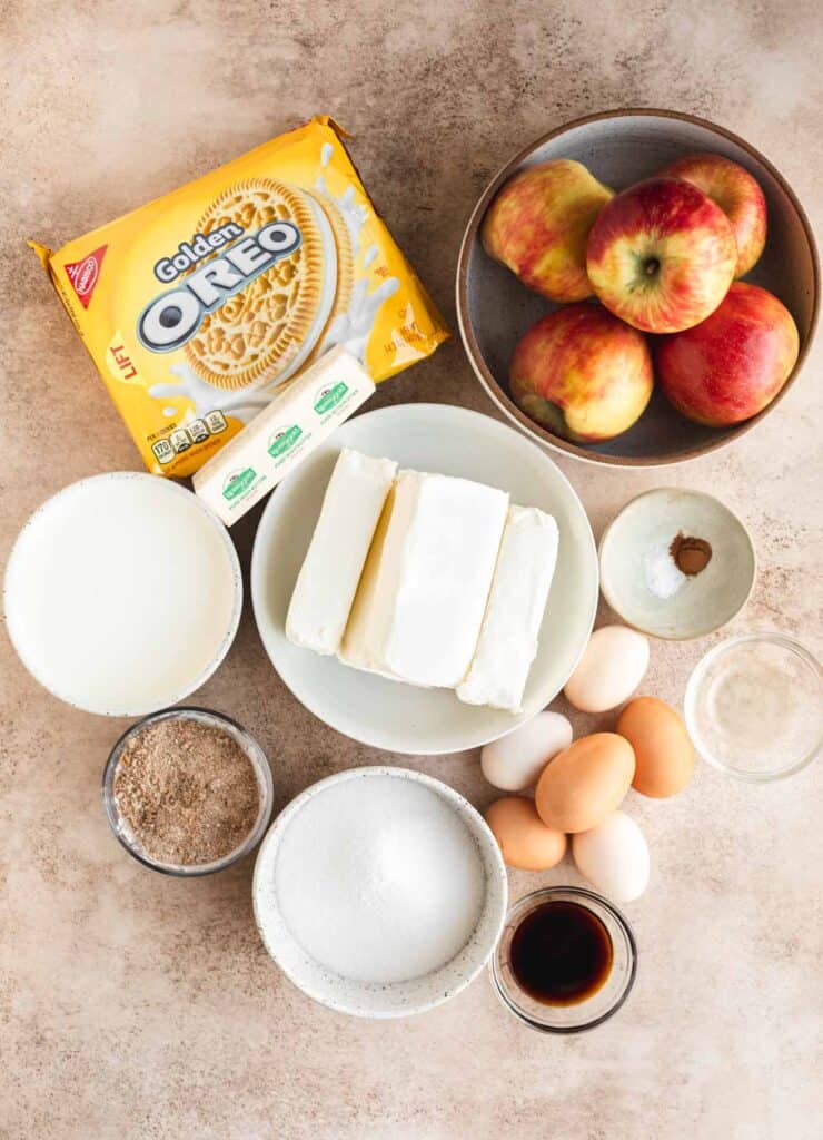 A package of Golden Oreos, a stick of butter, honey crisp apples in a bowl, salt, spices, eggs, vanilla, sugar, brown sugar, cream, sour cream and cream cheese in a bowl.