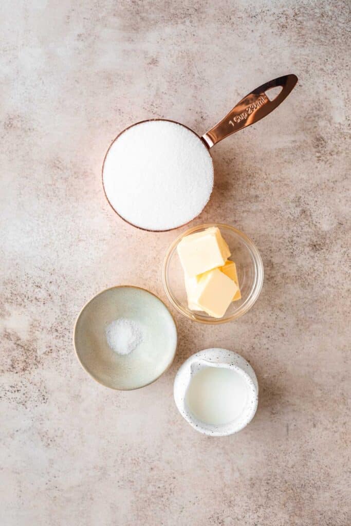 Granulated sugar in a copper measuring cup, cubed butter, salt and heavy cream in a small white pouring dish for the caramel sauce.