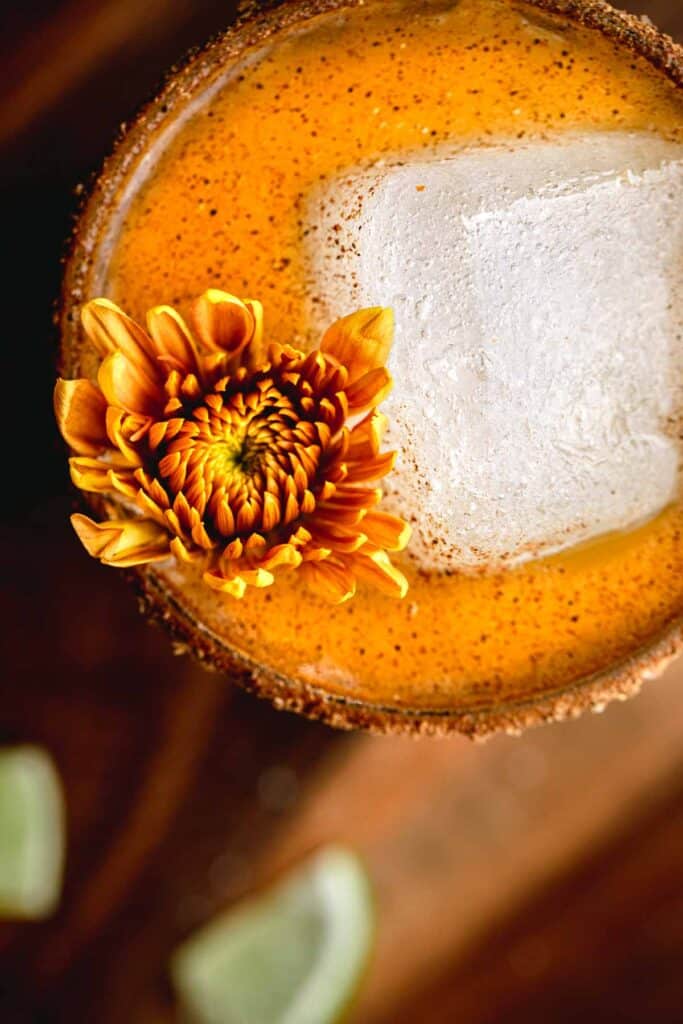 A yellow and orange mum flower in the rocks glass next to a large square chunk of ice in an orange margarita on a wooden board.