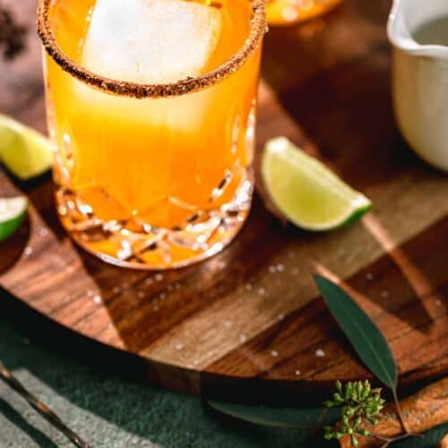 Pumpkin Spice Margaritas on a circular wooden serving tray with pumpkin spice and salt rims. A cocktail spoon, cinnamon stick and lime wedges to the side.