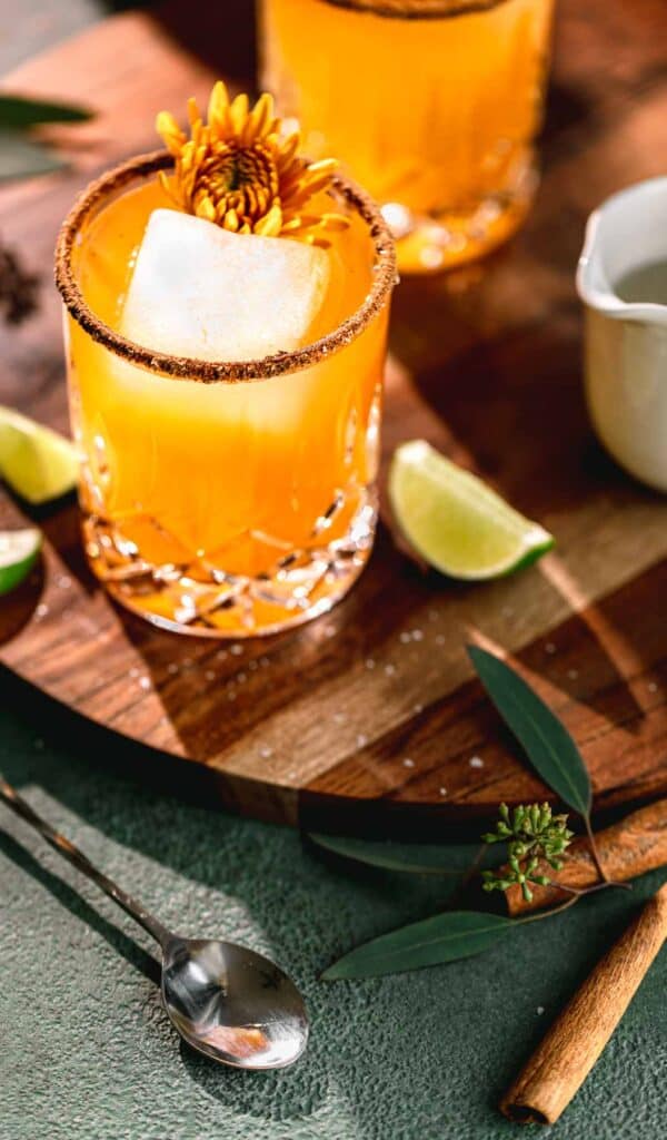 Pumpkin Spice Margaritas on a circular wooden serving tray with pumpkin spice and salt rims. A cocktail spoon, cinnamon stick and lime wedges to the side.